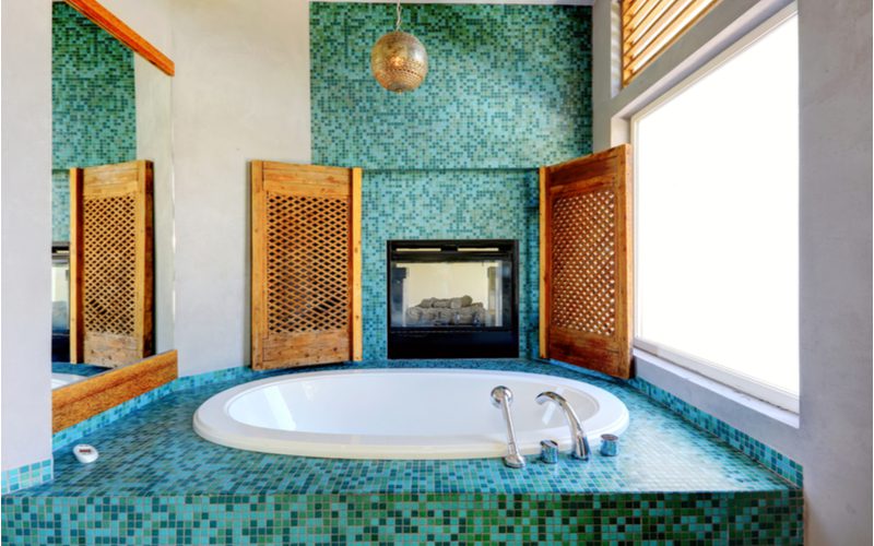 Идея бирюзового и зеленого мозаичного камина с камином рядом с большой ванной для купания, обрамленной ставнями из натурального дерева
